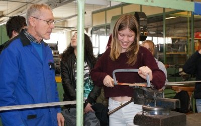 Das Bild zeigt eine Schülerin beim Sägen eines Metallstücks und einen Ausbilder